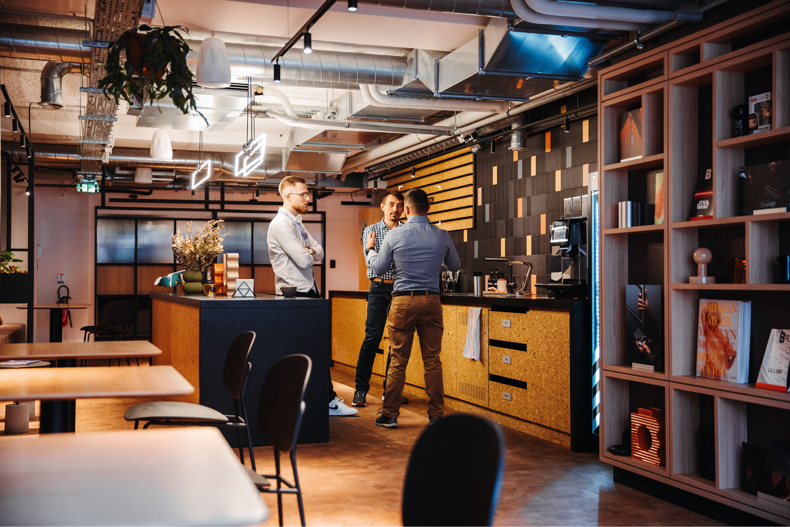 3 men stood in a small kitchen having a conversation, with shelving exposed industrial ceilings and tables and chairs to the left