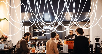 Three people queuing for a coffee with 2 baristas serving them, with long string lights hanging from the ceiling above them